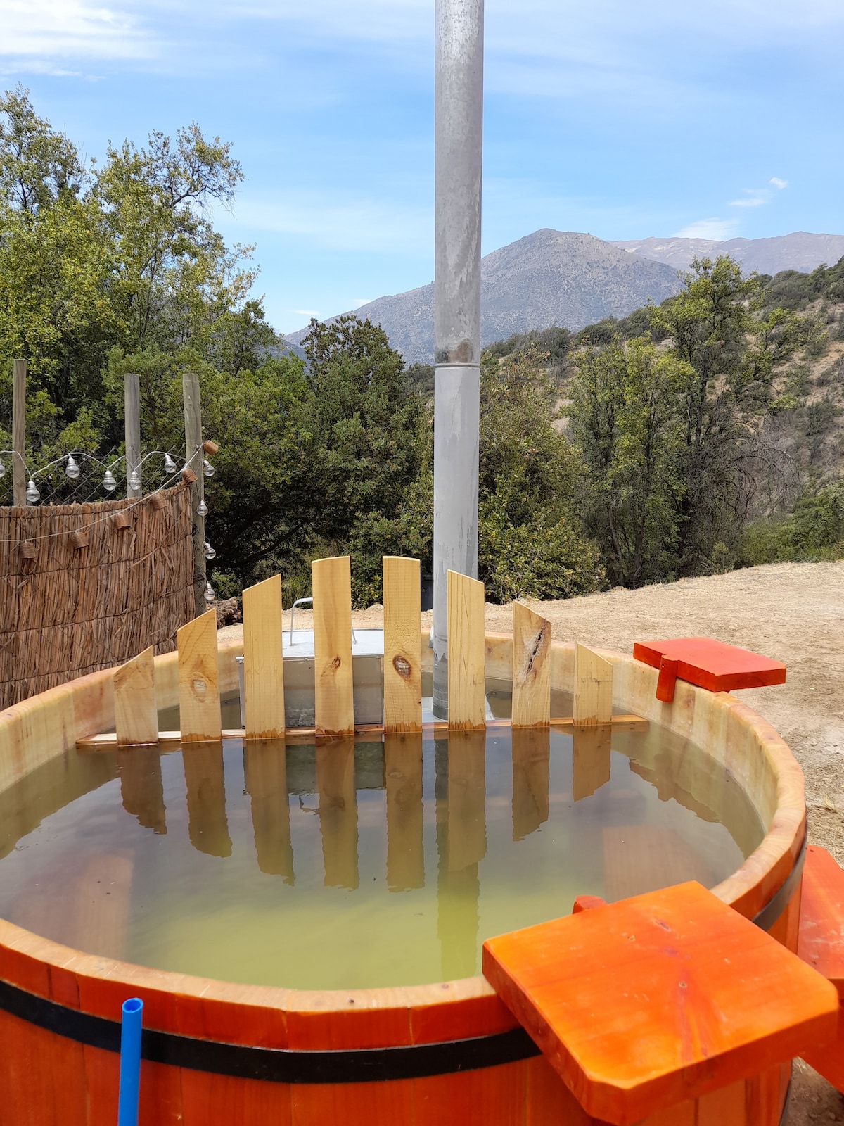 A rustic hot tub made of wood is set outdoors on a dirt path, surrounded by native trees and mountains in the background. Sunlight reflects off the water's surface, creating a serene atmosphere ideal for relaxation.