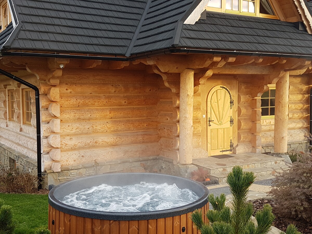 An outdoor jacuzzi is situated in a wooden enclosure, emitting steam in the cool air. The villa's log exterior, adorned with large windows and a wooden door, complements the natural landscape, creating a serene setting for relaxation.
