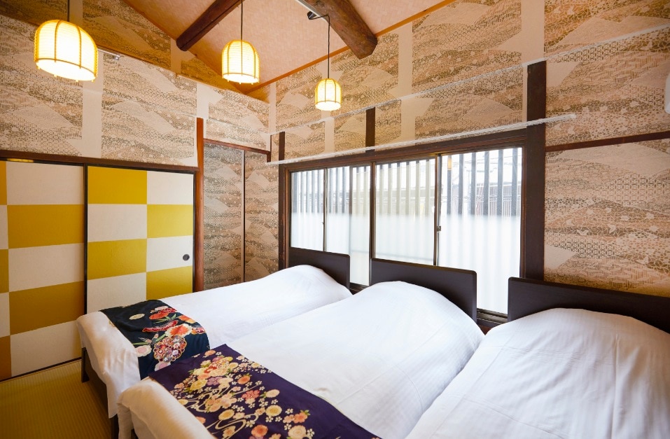 A bright bedroom is furnished with three spacious beds, each dressed in white linens. Decorative patterned walls add a touch of character, while natural light filters through the window, providing a welcoming ambiance. Traditional lighting fixtures enhance the room's serene atmosphere.