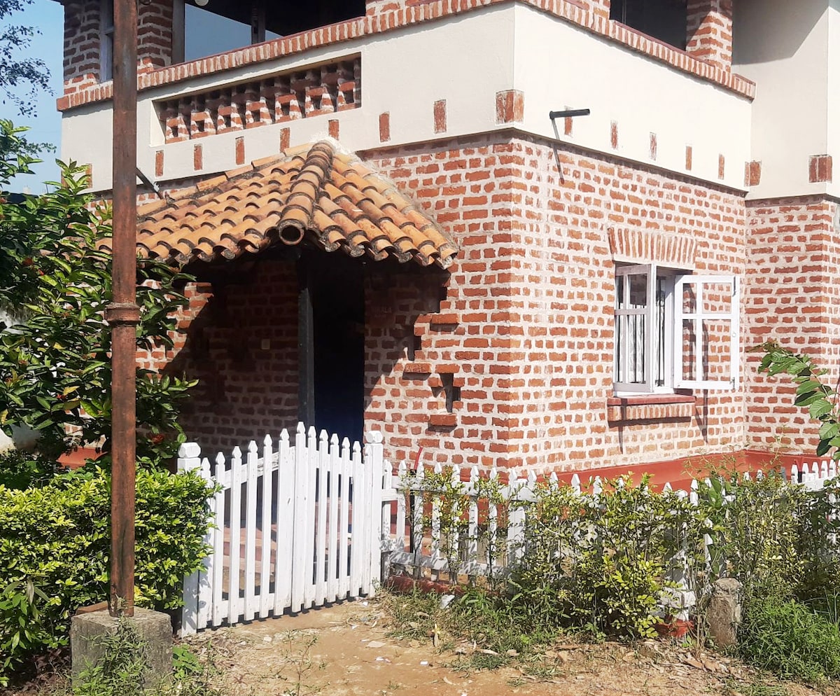The image features a rustic brick exterior of a bungalow, highlighted by a terracotta-tiled roof over the entrance. A white picket fence borders the property, complemented by green shrubs and small trees, creating a welcoming approach to the front door.