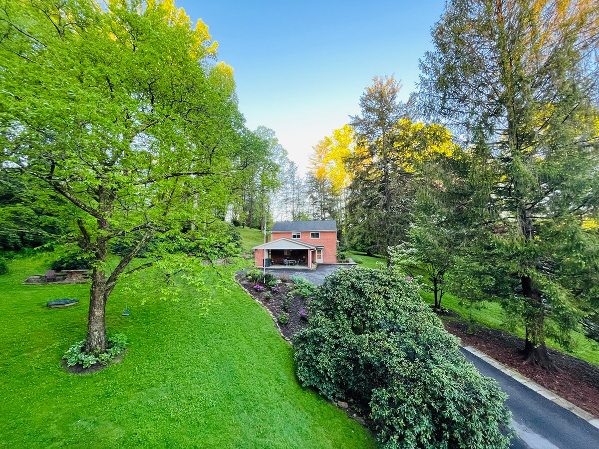 A spacious, well-maintained home is set against a backdrop of lush greenery and mature trees. The manicured lawn features a variety of plants, with a stone pathway leading to the entrance. The serene setting offers a peaceful retreat amidst nature.