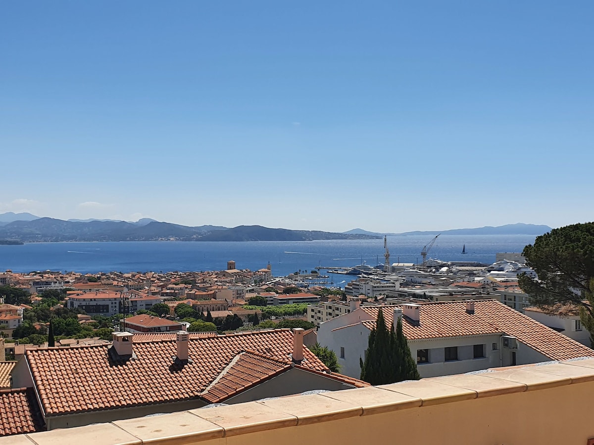 A panoramic view of the sea and distant mountains is captured from a terrace. The blue waters reflect the sky, while the town below is composed of terracotta rooftops. The harbor is visible, along with various vessels, contributing to the scenic landscape.