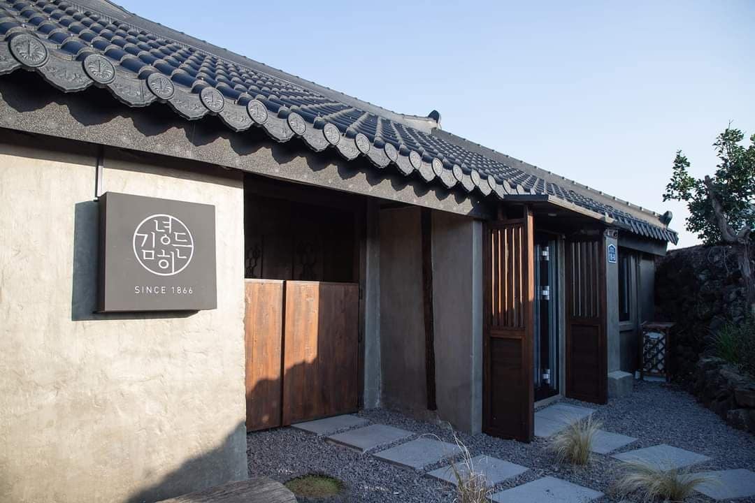 The exterior of a historic stone house reflects traditional Hanok architecture. A sign indicating 'Since 1866' is visible beside the entrance. Wooden doors partially open, revealing a pathway lined with stepping stones and small vegetation, leading to the houseβs inviting entrance.