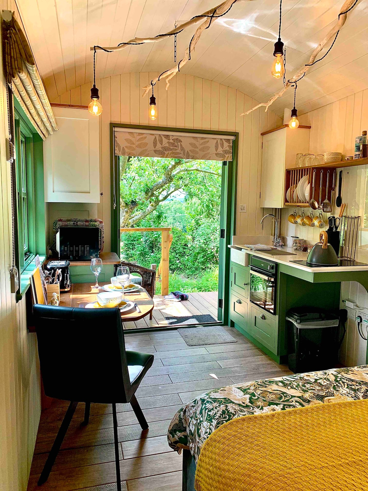 The interior of the shepherd's hut features a well-equipped kitchenette with a gas oven, sink, and dining area with a table set for four. Natural light floods in through the door leading to an outdoor space, while soft lighting hangs from the ceiling.