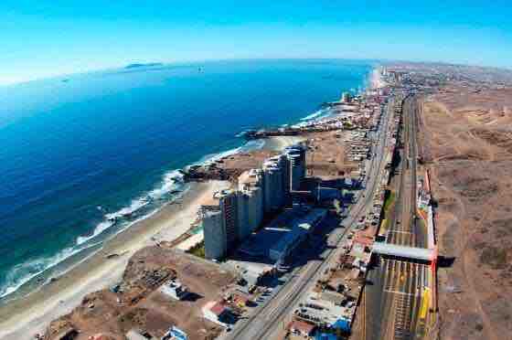 An aerial view captures a long stretch of coastline, where the ocean meets the sandy beach. The beachfront is lined with various buildings, with a road running parallel to the shore. The background features distant waves and a clear blue sky.