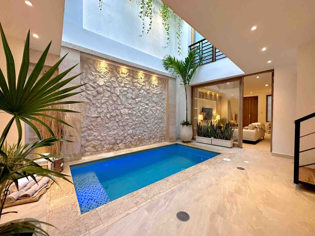 A serene indoor pool area features a small pool surrounded by natural stone walls and lush greenery. Sliding glass doors open to a bright living space, allowing natural light to fill the room. A cozy seating area is visible in the background, enhancing the atmosphere.