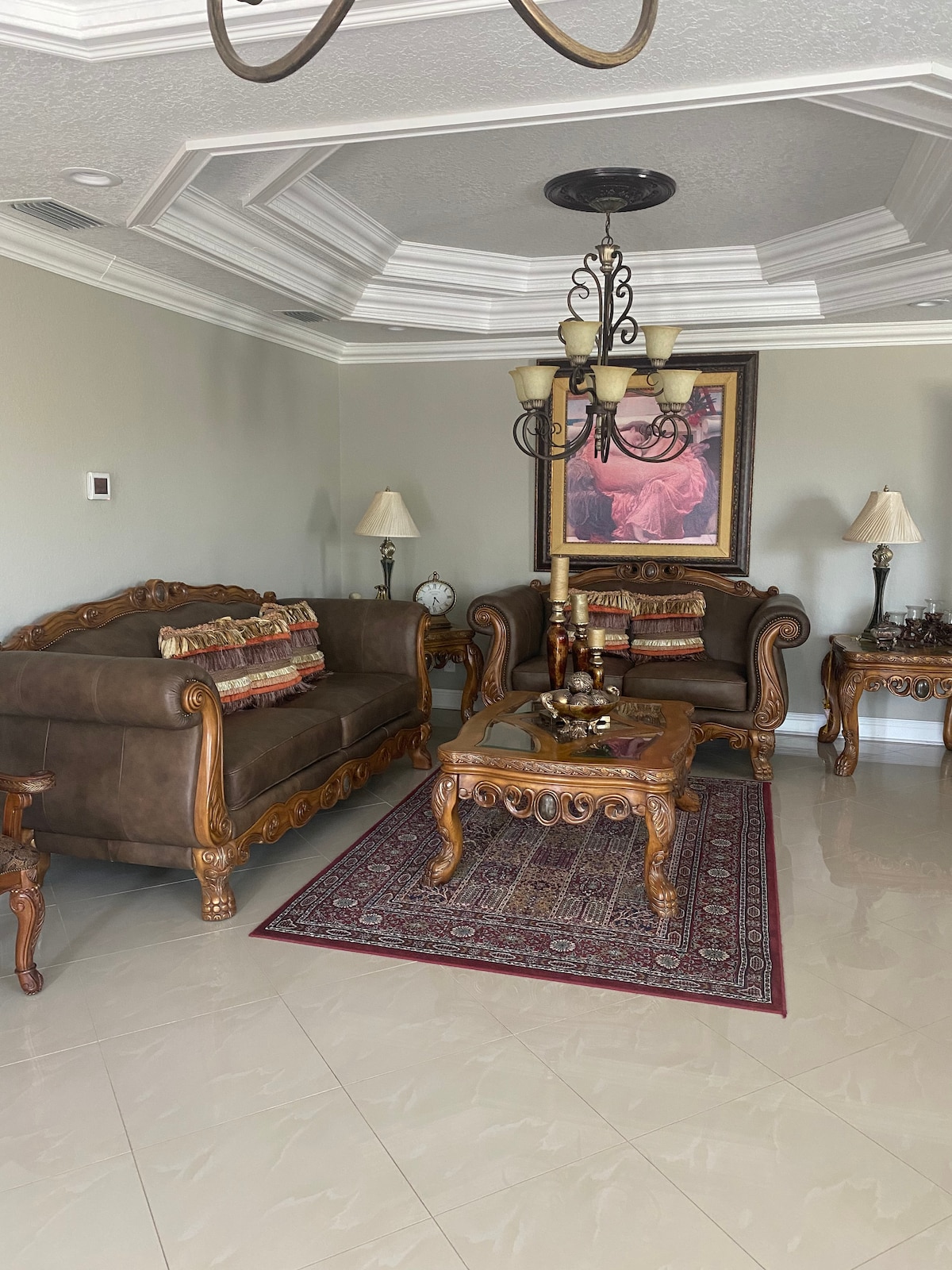 A living area features ornate furniture with dark upholstery, complemented by a decorative area rug. Elegant lamps are placed on side tables, and a large framed artwork adorns the wall above a sofa. The ceiling showcases detailed molding, adding a touch of sophistication.