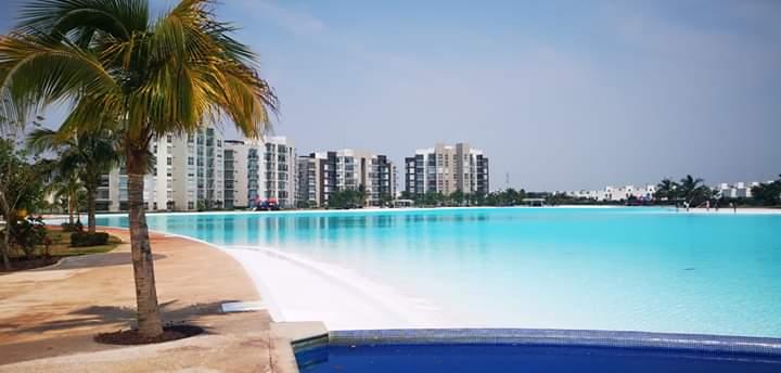 A tranquil artificial lagoon extends alongside modern buildings, reflecting bright blue waters. Lush palm trees provide shade by the water’s edge, while the surrounding area features sandy pathways and green landscaping, enhancing the accessibility of the space for leisure and relaxation.