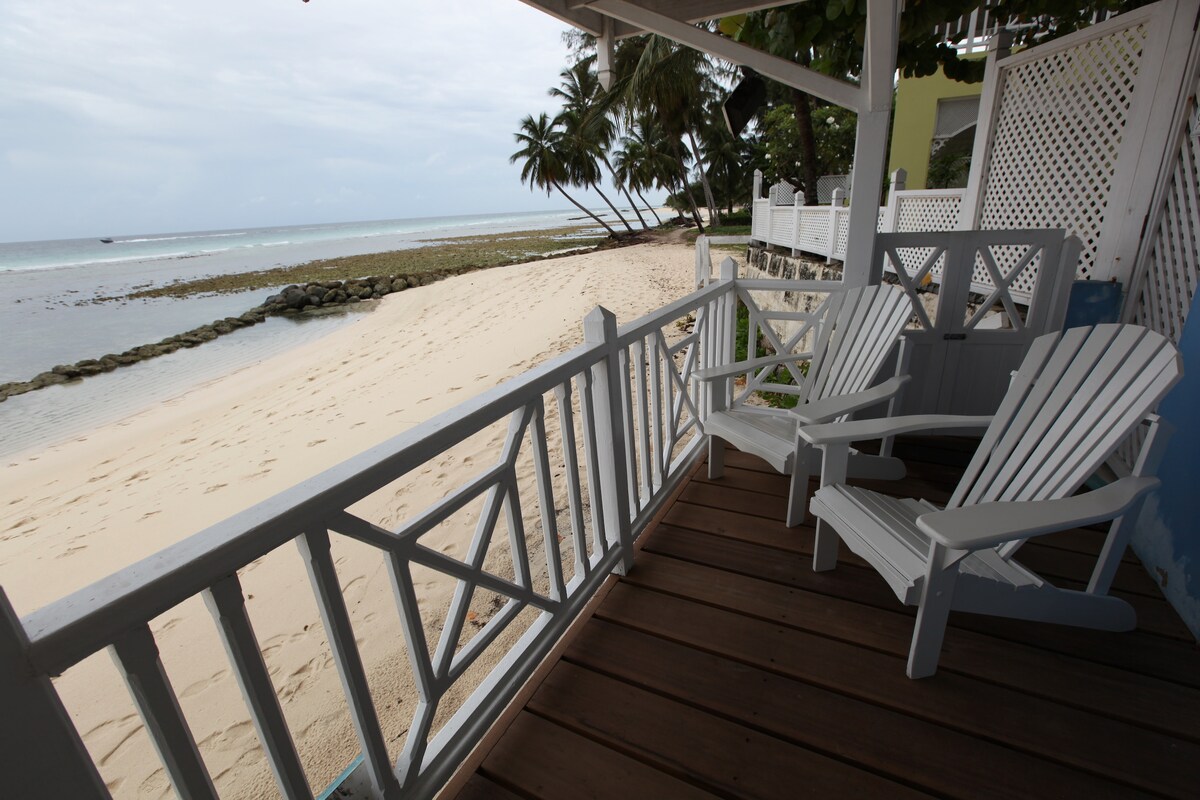 A charming verandah features two white Adirondack chairs positioned invitingly, overlooking the sandy beach and palm trees swaying in the breeze. The sea glistens in the background against a calm sky, enhancing the tranquil coastal setting.