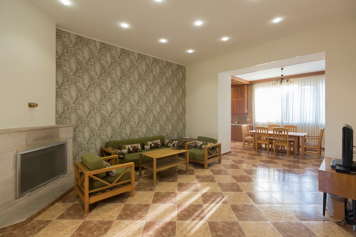 A spacious living area is shown with light-colored walls and a patterned accent wall. Comfortable seating arrangements consist of green sofas and wooden coffee tables. Behind, a dining space with a wooden table and chairs is visible, illuminated by natural light from large windows.
