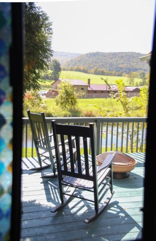 Two rocking chairs are positioned on a wooden deck, overlooking a scenic view of lush fields and rolling hills. A small fire pit is placed nearby, with sunlight casting a warm glow over the peaceful outdoor space.