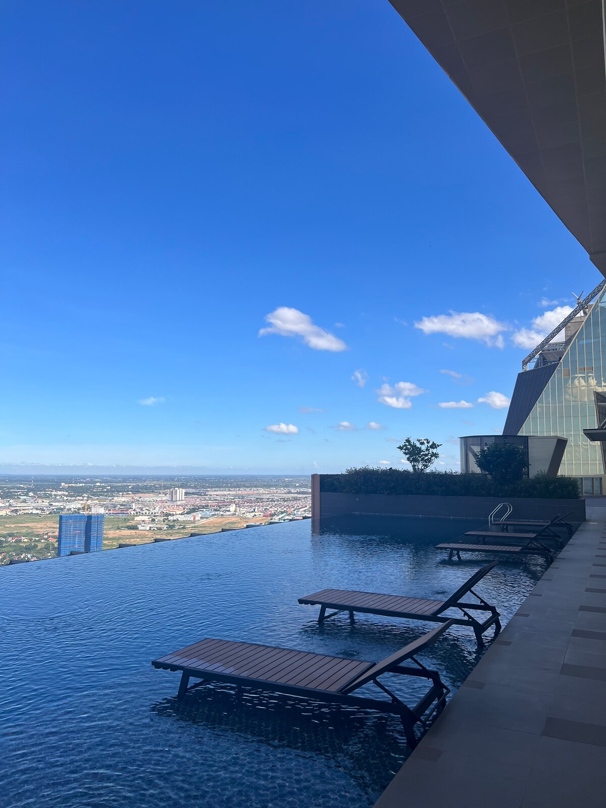 An infinity pool extends along the edge of a high-rise building, reflecting the bright blue sky and offering panoramic city views. Wooden loungers are positioned beside the pool, providing space for relaxation. Lush greenery is visible along the building's edge, enhancing the tranquil setting.