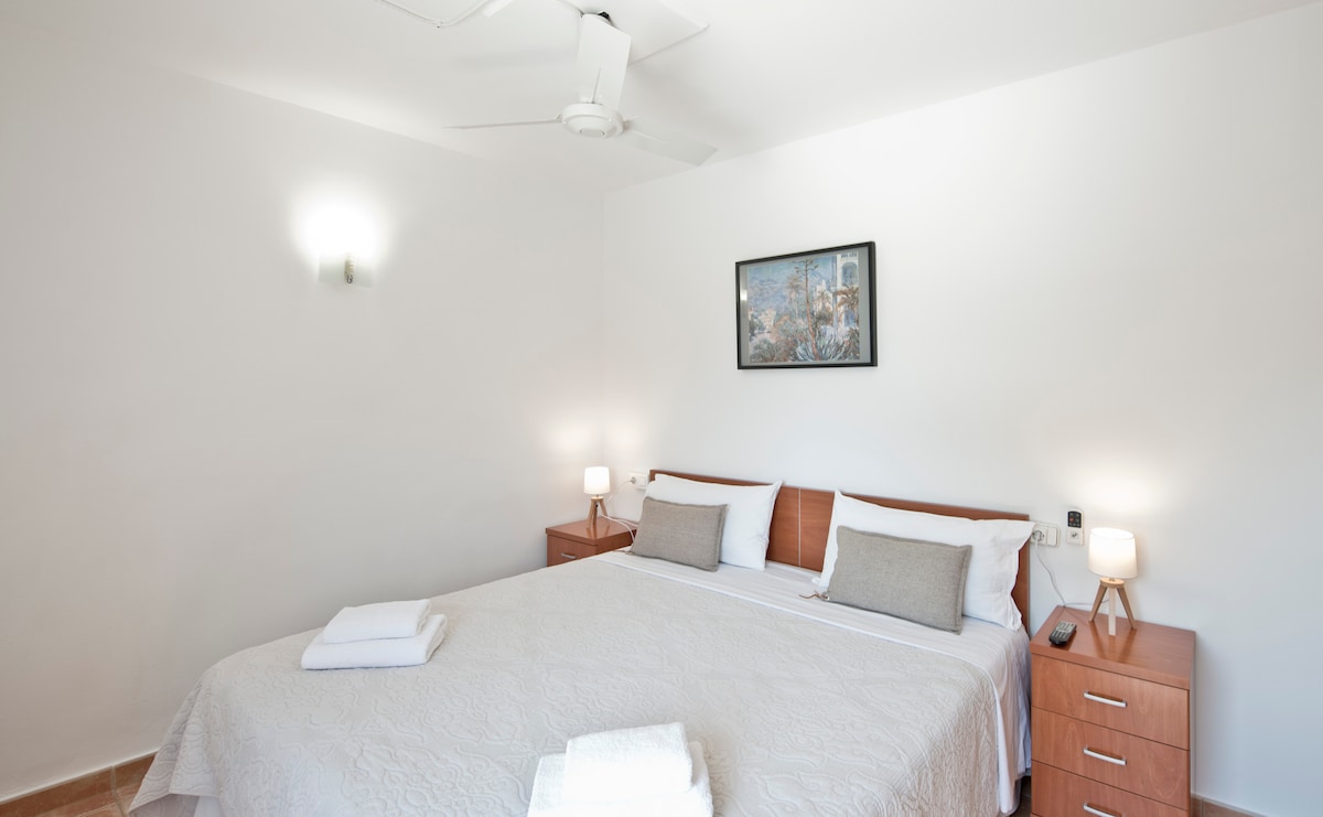 A bright bedroom features a spacious bed adorned with a light-colored bedspread. Two bedside tables with lamps provide soft lighting. A ceiling fan contributes to air circulation. A framed artwork hangs above the bed, complementing the minimalist decor.