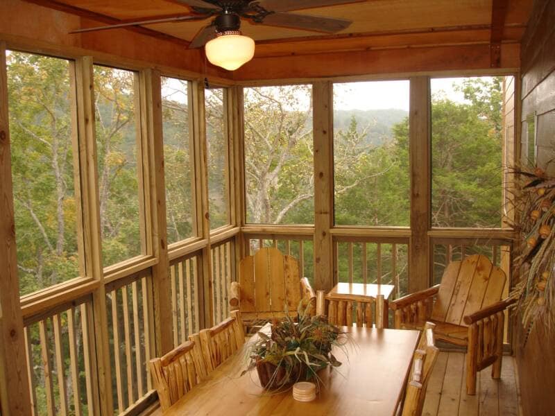 A screened porch features rustic wooden furniture, including two chairs and a table. Natural light filters through large windows that provide views of surrounding trees and the hillside, creating a serene and cozy atmosphere.