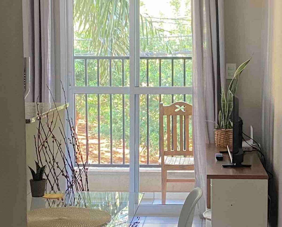 A sunlit interior features large glass doors opening to a balcony with views of greenery. A wooden chair sits beside a desk, complemented by a small plant and a television on the surface. Sheer curtains allow natural light to fill the space.