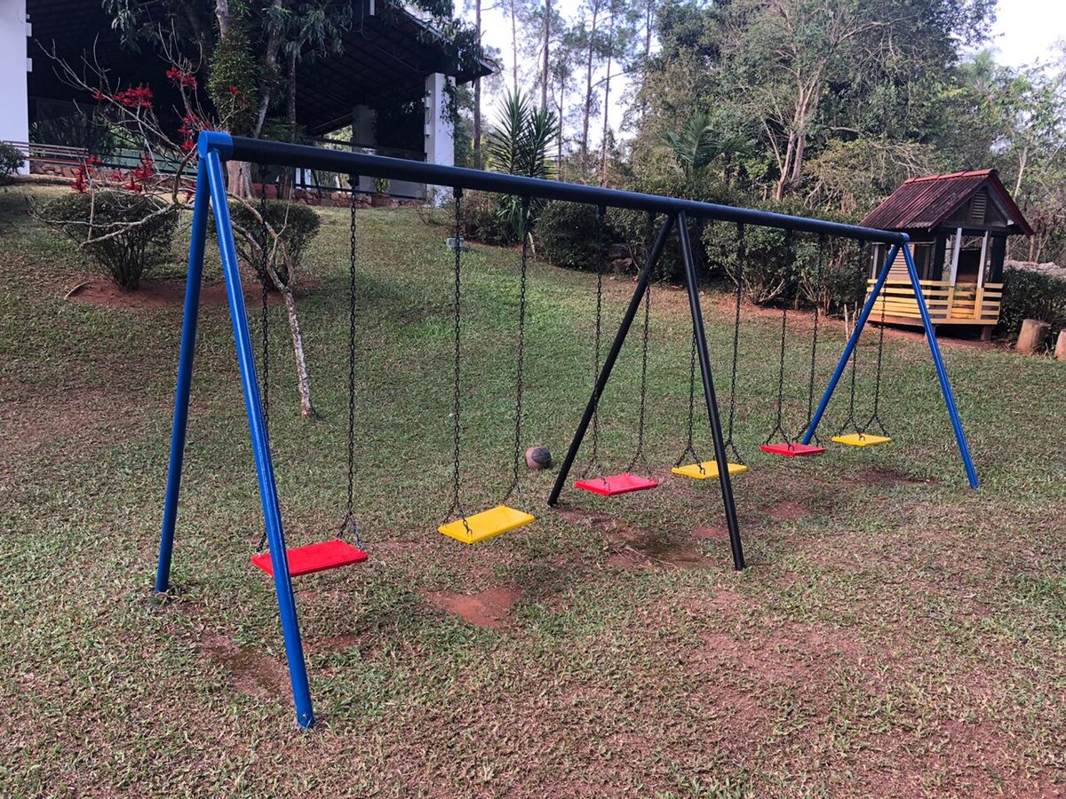 A playground area features a swing set with five seats, alternating between red and yellow colors. The swings hang from a sturdy black frame, surrounded by lush green grass and trees. A small wooden playhouse can be seen in the background.