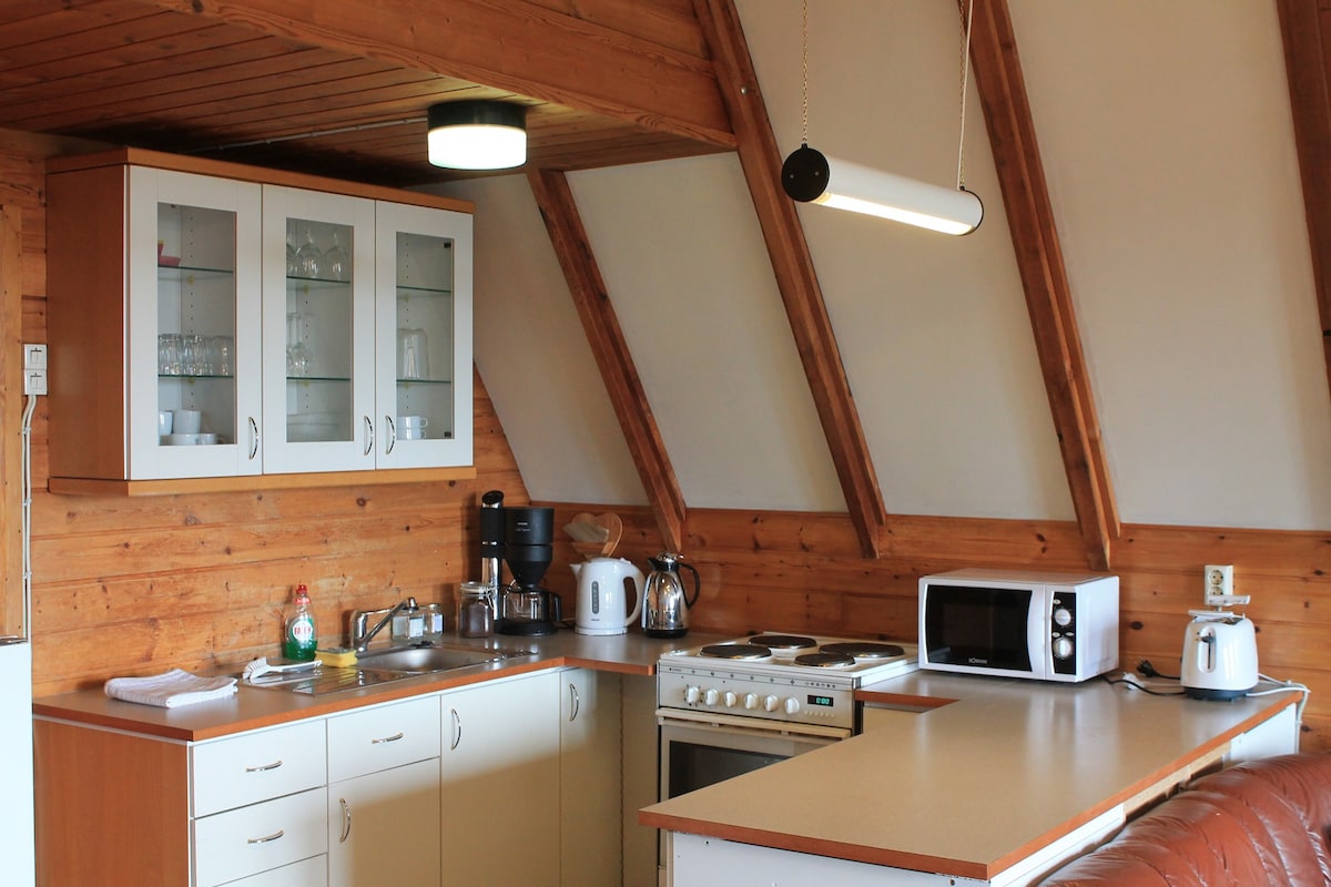 The kitchenette features wooden walls and a sloped ceiling, providing a cozy atmosphere. Appliances include a microwave, stove, and coffee maker. Cabinets display glassware. A counter offers a spacious work area, complemented by a modern light fixture above.
