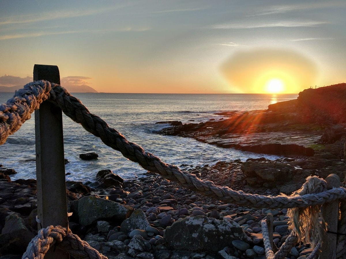 The image captures a serene sunset over the ocean, with the sun dipping below the horizon and casting warm hues across the sky. A rocky shoreline is visible in the foreground, adorned with a textured rope railing that adds a rustic element to the coastal scene.