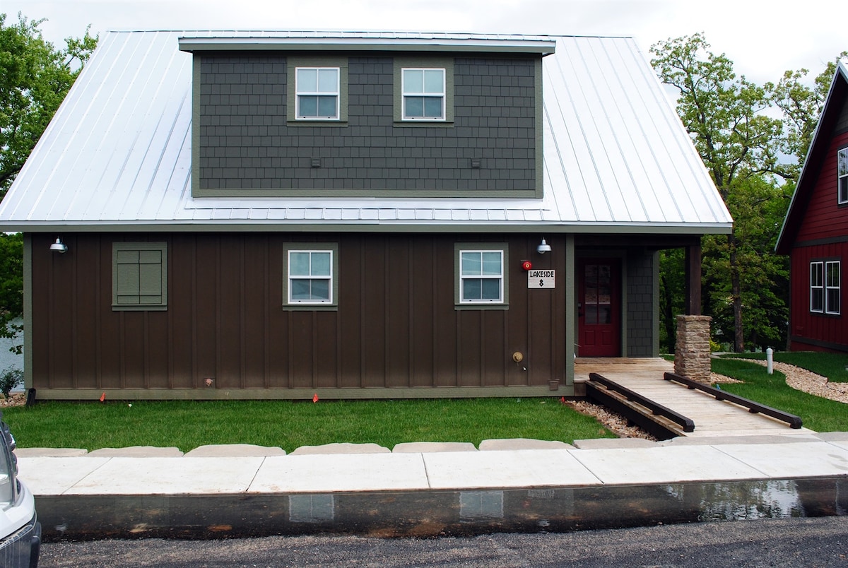 A modern cabin exterior is depicted, featuring a brown wooden facade complemented by a metal roof. A covered entrance is highlighted, with a concrete walkway leading to the door. Lush green grass surrounds the cabin, enhancing the natural setting.