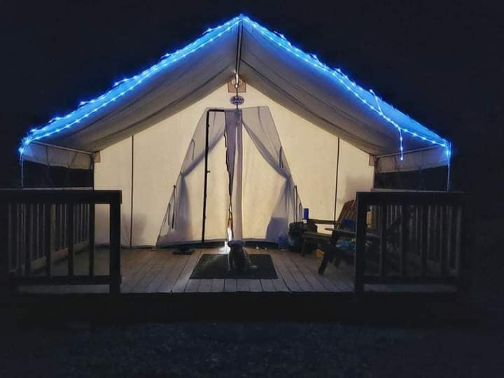 A spacious glamping tent is illuminated by soft blue lighting at night. The entrance is framed with sheer curtains, revealing a welcoming interior. A wooden deck features seating, and a small rug is centered at the entryway.