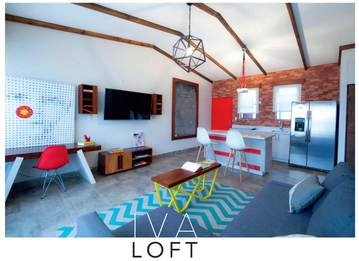 The living area features a modern sofa and a 55-inch LED TV mounted on a textured wall. A stylish coffee table sits atop a patterned rug. A workspace with colorful chairs and a fully-equipped kitchen is visible in the background, illuminated by natural light from large windows.