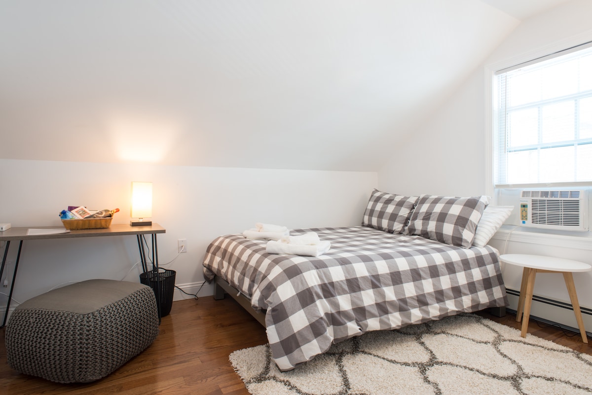 A cozy private room features a full-size bed with a plaid duvet cover and plush pillows. A small desk is set nearby, accompanied by a stylish stool and a decorative basket. Natural light floods the space through a window, while a soft area rug adds warmth to the wooden floor.