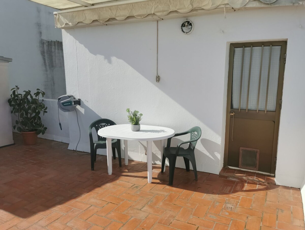 A small outdoor terrace features a round white table surrounded by two chairs, one black and one green. A potted plant adds a touch of greenery, while the wall is painted white. A door leads into the interior space.