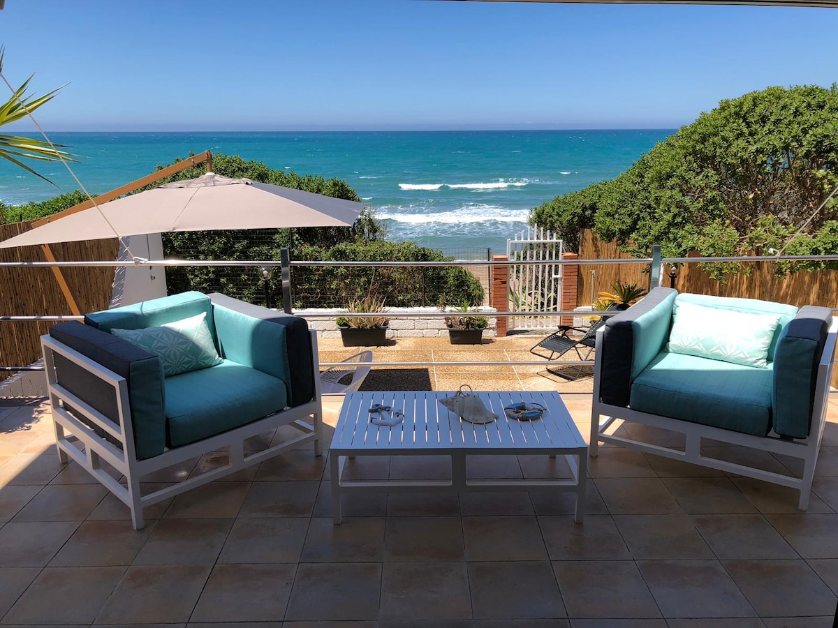 A comfortable outdoor seating area offers views of the ocean, featuring two light blue armchairs and a white coffee table. Sunlight reflects off the water, while lush greenery and a patio umbrella complete the serene environment.
