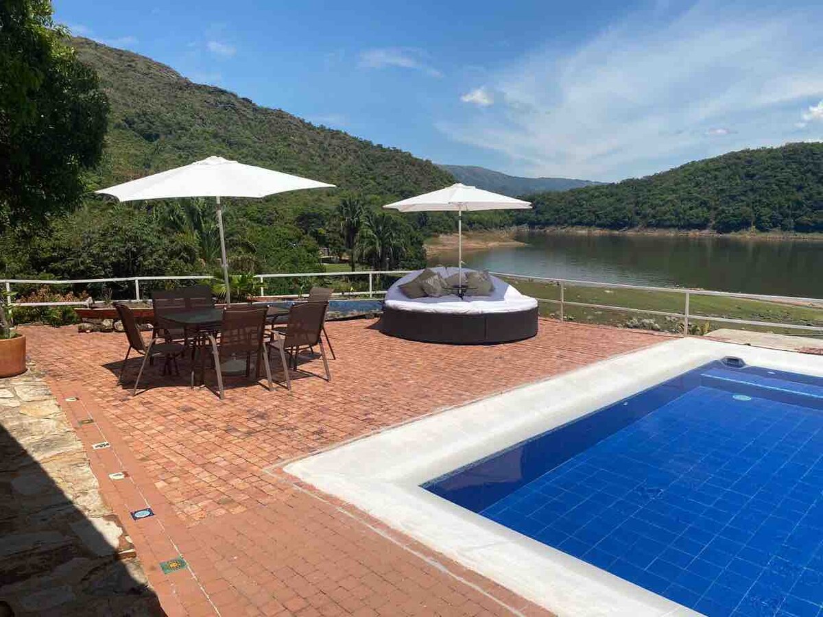A serene outdoor terrace is highlighted by a spacious pool with clear blue water and surrounded by inviting seating areas. Shade is provided by large umbrellas, while views of lush hills and peaceful waters create a tranquil atmosphere.