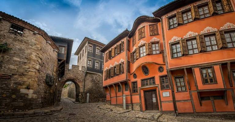A historic street scene showcases traditional Bulgarian architecture, featuring colorful buildings with intricate wooden window frames. A cobblestone path winds between the structures, with a stone wall partially visible in the background. The sky above is bright, contributing to a charming ambiance.