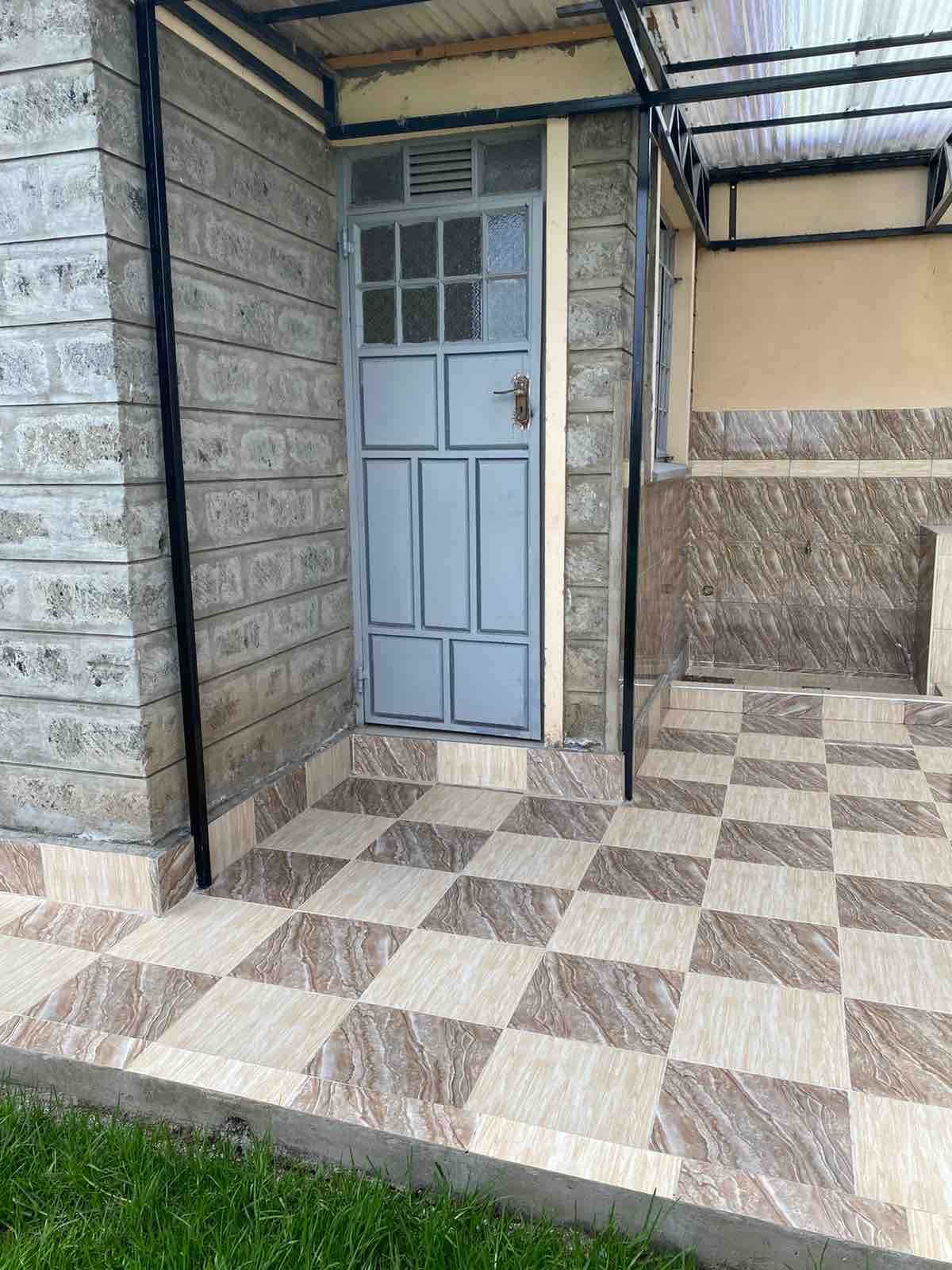 The entrance features a light blue door with a single handle, framed by a patterned stone wall. The spacious patio showcases a checkerboard tile design in browns and creams. Green grass borders the tiled area, creating a natural contrast.