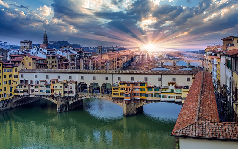 A panoramic view showcases the iconic Ponte Vecchio illuminated by a vibrant sunset. The Arno River reflects the colors of the sky, while historic buildings and bridges frame the scene, capturing the rich architectural heritage of Florence.