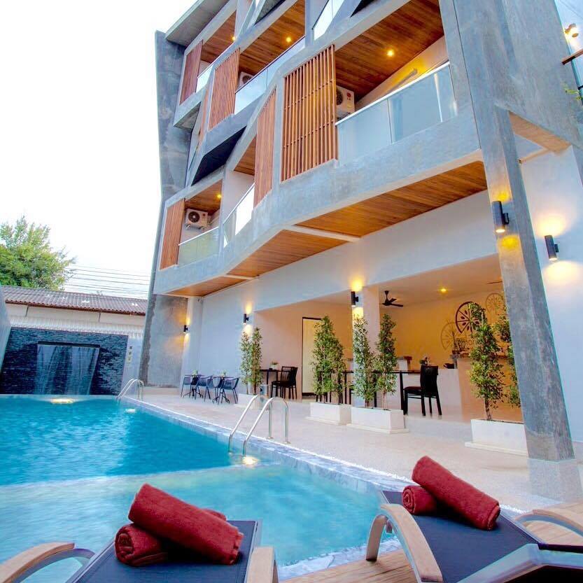 The building features a contemporary design with large balconies and a poolside area. Sun loungers are positioned around the inviting pool, and a relaxed atmosphere is created by the surrounding greenery. The entrance to the lobby is visible, enhancing accessibility.