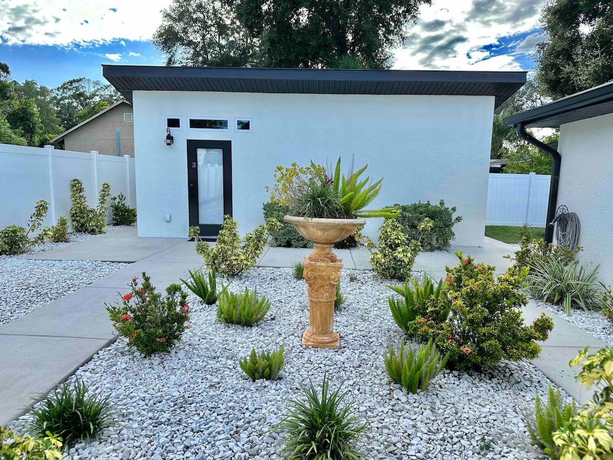 A well-kept outdoor area is visible, featuring a decorative stone fountain surrounded by gravel and various plants. A manicured garden with shrubs and flowers enhances the entryway. The home's entrance is framed by a modern door with an inviting window beside it.