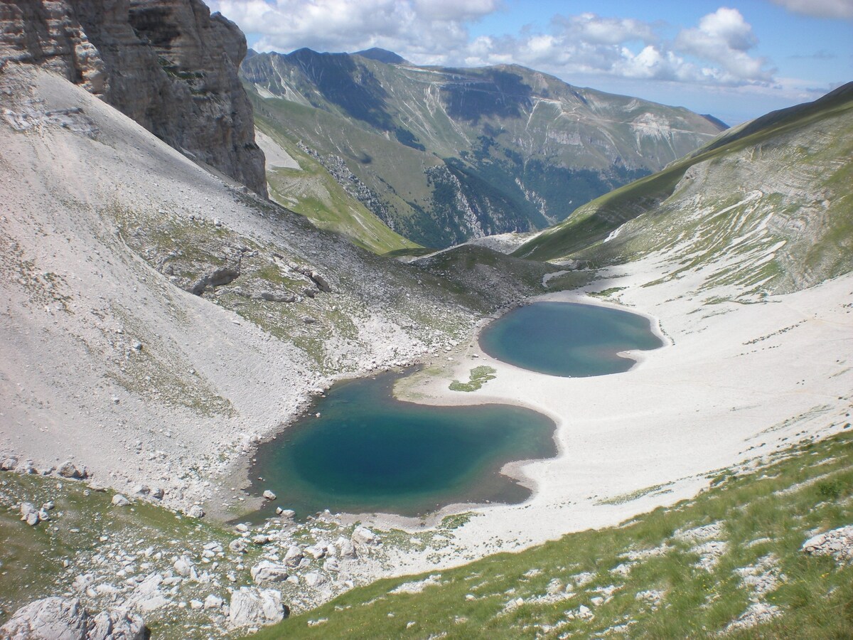 Two emerald-colored lakes appear nestled within a mountainous landscape, surrounded by sloping hills and rocky terrain. A light sandy area borders the lakes, contributing to a serene natural environment. Bright clouds drift across a blue sky, enhancing the peaceful ambiance.