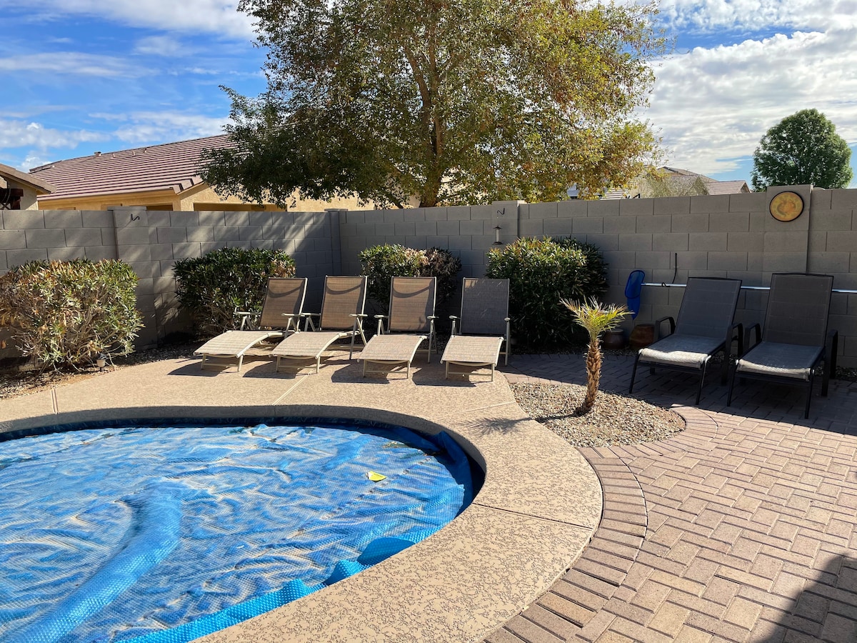 An outdoor area features a pool covered with a blue tarp, surrounded by a tiled patio. Several lounge chairs are positioned along the edge, with lush shrubs and a small palm tree providing greenery. The sky is partly cloudy, offering a pleasant atmosphere.