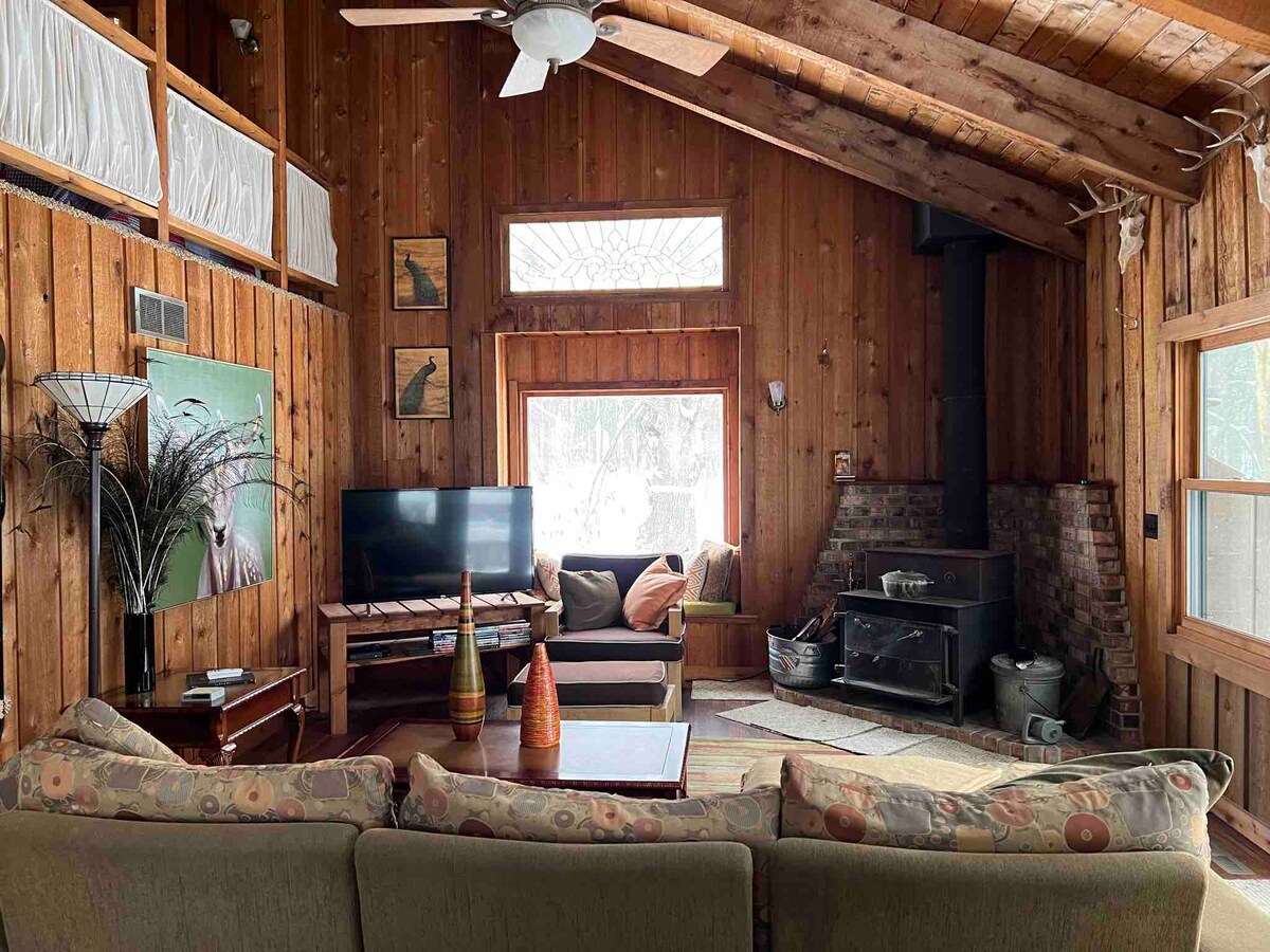 A cozy living area features wooden paneling and beamed ceilings, with a large sofa facing a television. A wood-burning stove is situated in the corner, alongside a rustic brick hearth. Natural light floods in through large windows, enhancing the warm atmosphere.