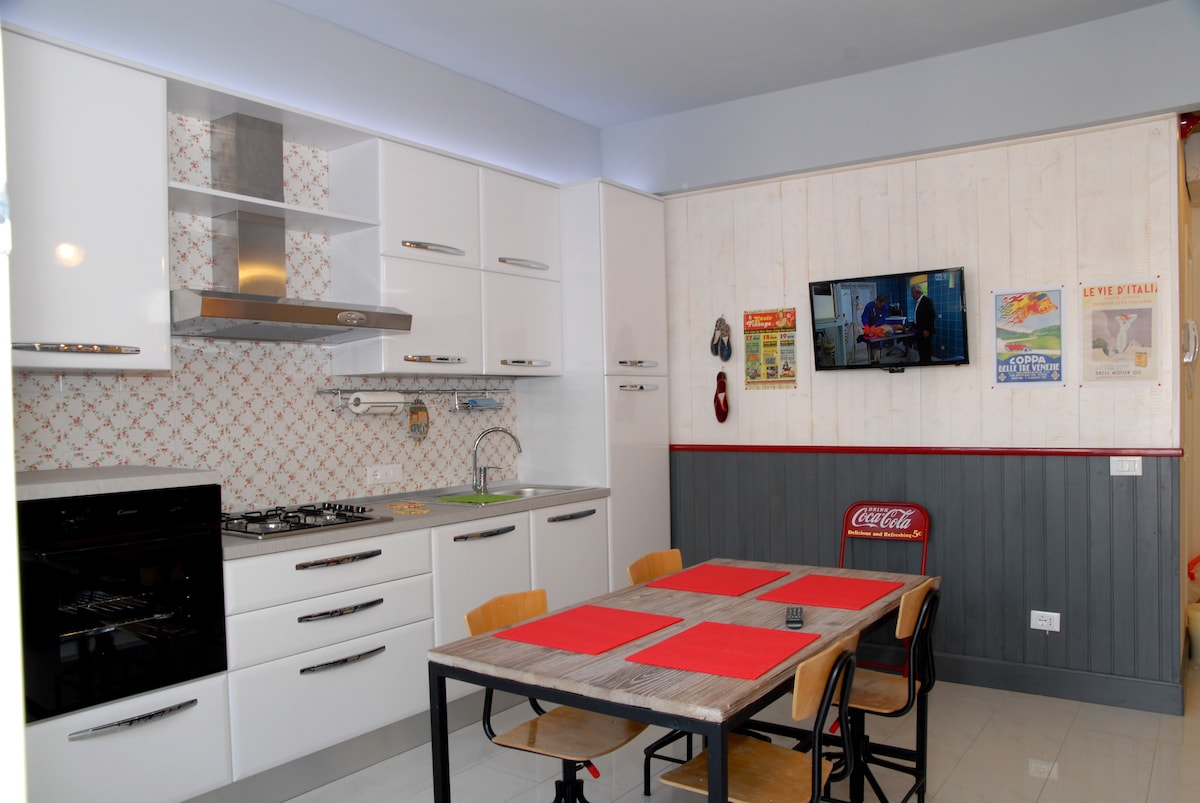 A modern kitchen is equipped with sleek white cabinets and a stylish backsplash. A dining table with six wooden chairs is set with red placemats. Contemporary appliances, including a gas stove and a wall-mounted TV, complete the space, offering both functionality and comfort.