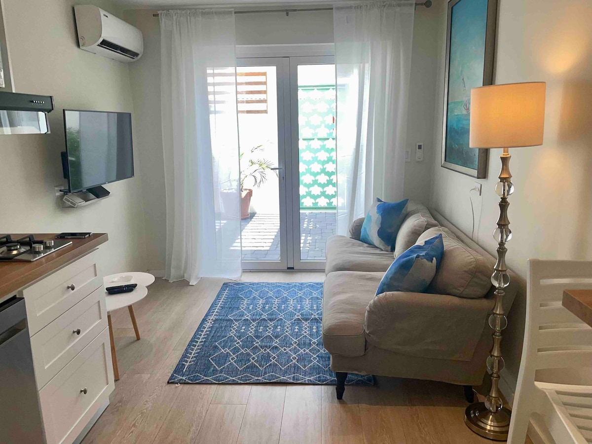 A cozy living space features a light-colored couch adorned with blue cushions. A small table and a television are positioned nearby. Natural light filters through sheer curtains, illuminating the room. A sliding glass door leads to a small outdoor area, visible through patterned privacy screens.