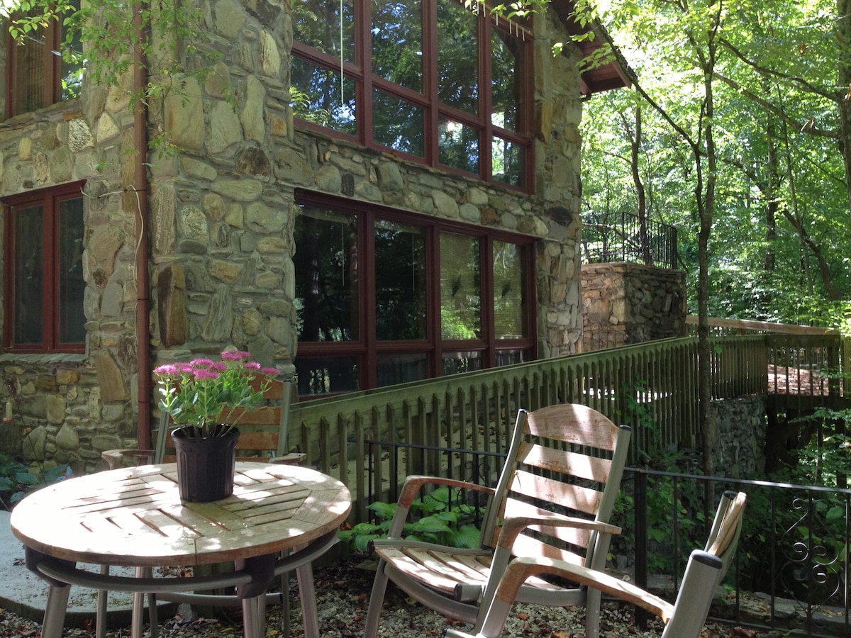 An outdoor seating area features a round wooden table with a potted plant and two wooden chairs. The background showcases a stone building with large windows framed by trees, enhancing the connection to nature.
