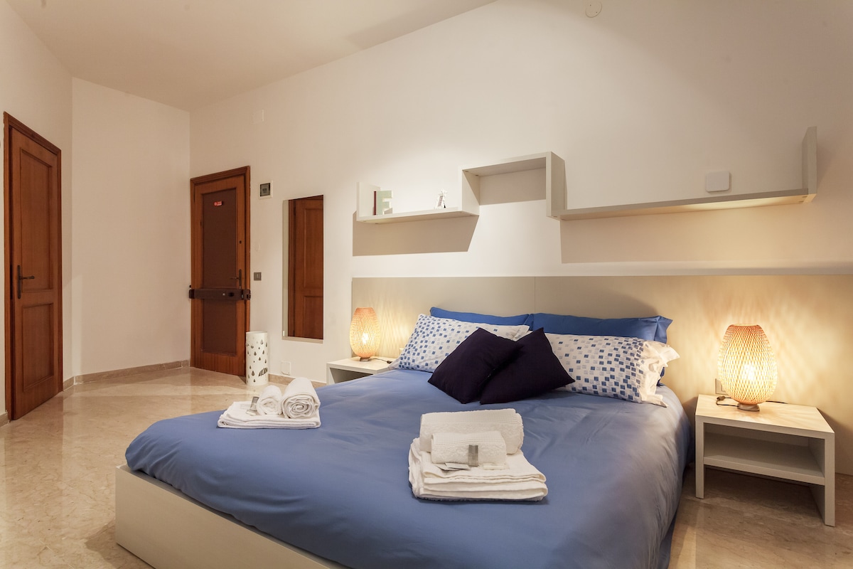 A tranquil bedroom is highlighted by soft lighting from two decorative lamps positioned on either side of a spacious bed. The bed is made with crisp linens in calming blue tones, complemented by decorative pillows. Shelving above the bed adds a modern touch, while a neutral wall enhances the serene atmosphere.