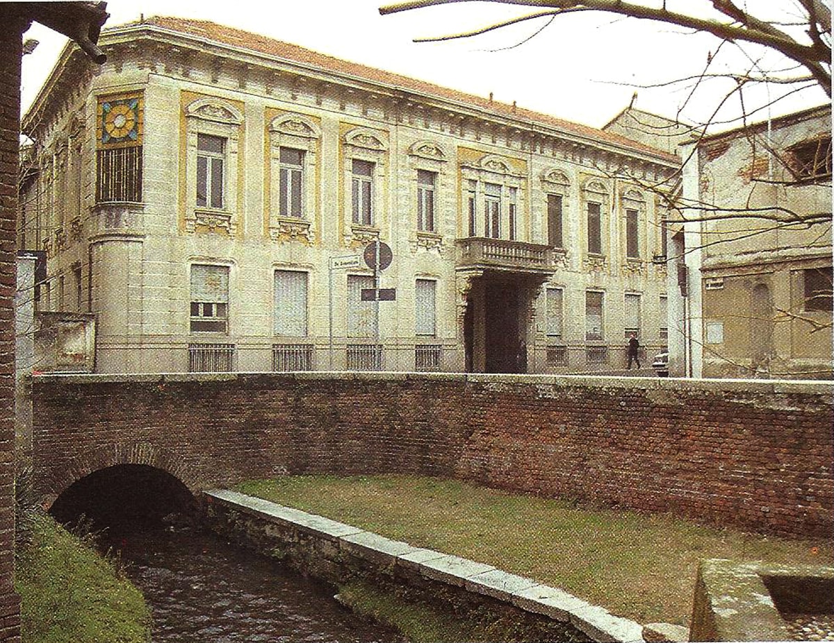 A historic building features decorative façades with ornate designs and a central balcony. A small bridge spans a flowing stream in the foreground, complemented by a grassy area. The scene is framed by bare tree branches, highlighting the structure's timeless character.