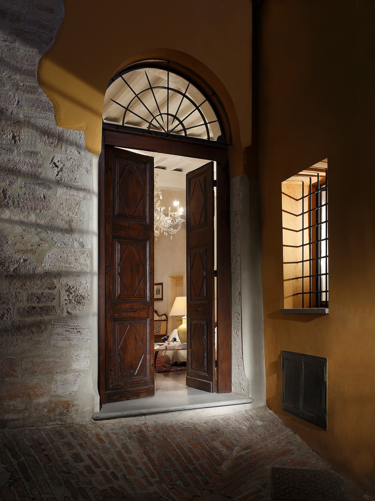 An inviting entrance features tall wooden doors set in a stone archway, allowing glimpses of the living area beyond. Soft light streams in, illuminating the warm walls. A decorative wrought-iron window is visible to the right, adding a touch of architectural detail.