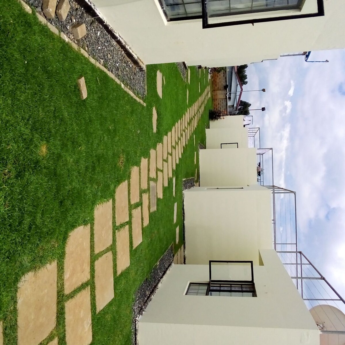A well-maintained outdoor area features a pathway of light-colored stones, leading through a grassy lawn. Neatly trimmed grass is bordered by gravel, with white walls of adjacent buildings visible in the background. Natural light is present, creating an open and inviting space.