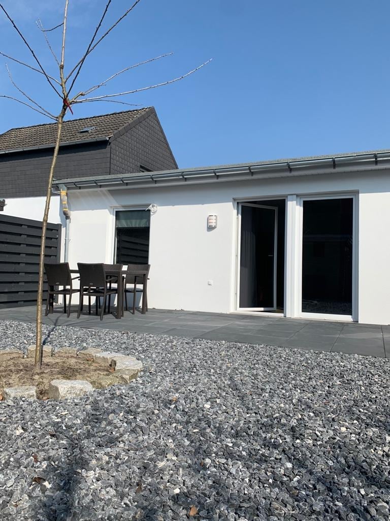 An outdoor area is shown, featuring a stone patio with a small tree planted in the center. A set of dark outdoor furniture, including a table and several chairs, is visible. The exterior walls of the property are white, complemented by large glass doors leading into the interior.