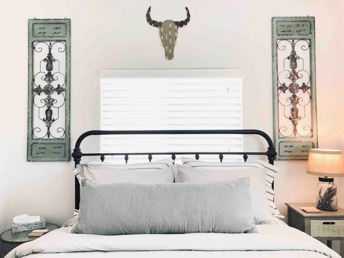 A calming sleeping area features a spacious bed with layered linens and a long decorative pillow. Two framed wall art pieces flank the headboard, complemented by a lamp on a nightstand. Natural light filters through a partially covered window above the bed.