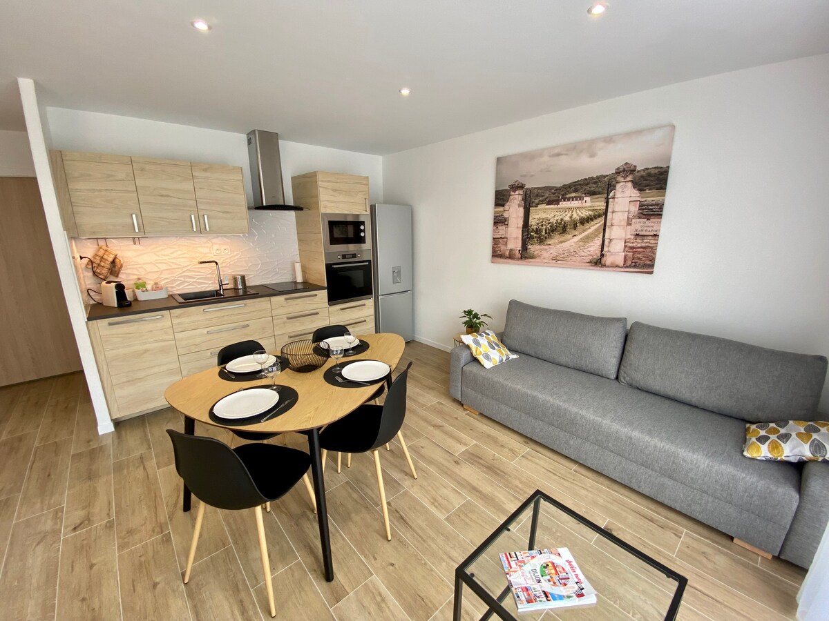 A modern living area features a wooden dining table set for four, accompanied by black chairs. A gray sofa sits against the wall, and a kitchen area with contemporary appliances is visible. Natural light brightens the space through a large window, enhancing the neutral tones.