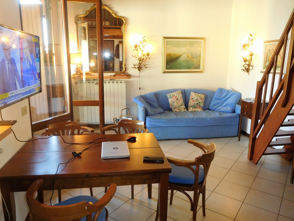 A living area features a blue sofa adorned with floral pillows, alongside a wooden table and chairs. A television is mounted for viewing, and light is provided by two wall sconces. Large windows allow natural light, enhancing the overall welcoming space.