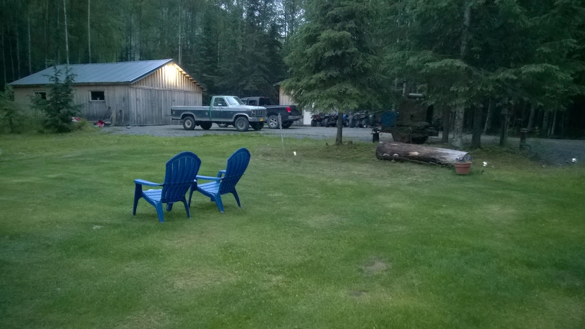 A grassy yard is visible, featuring two blue Adirondack chairs positioned on the lawn. In the background, a rustic cabin and a parked truck can be seen, surrounded by trees. A log and potted plant add to the outdoor setting.