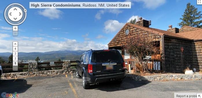 A spacious parking area is shown in front of a rustic-style condominium. A black SUV is parked in the foreground, with views of the surrounding mountains visible in the background. The building is constructed with wooden panels and features a stone accent.